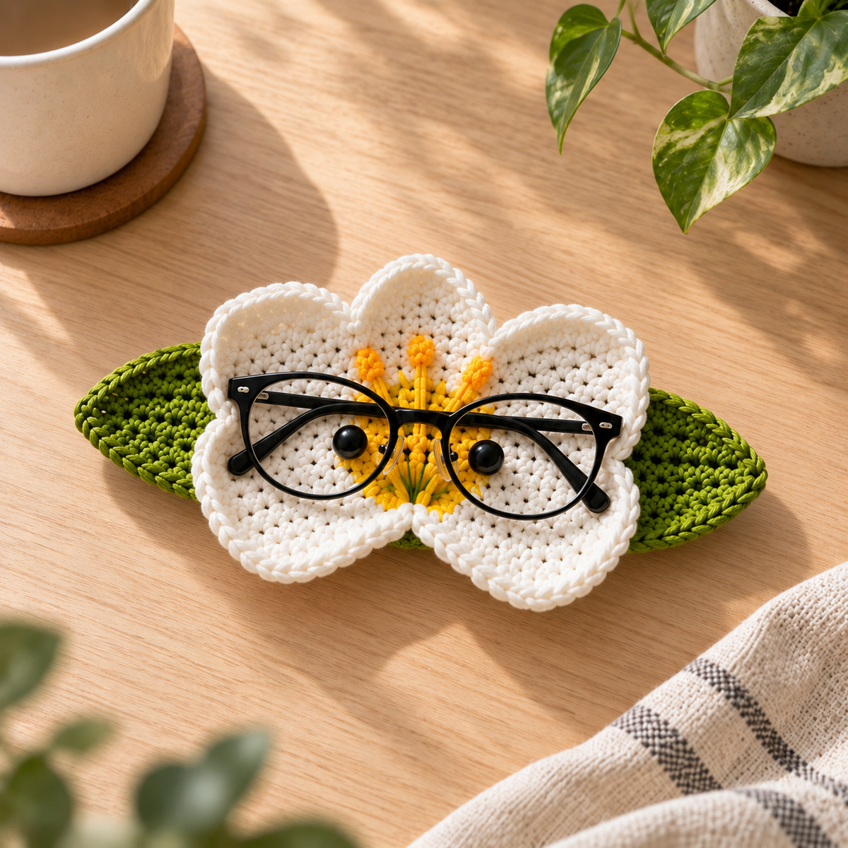 Handmade crochet glasses holder in a butterfly-inspired white and yellow floral design with green leaf accents, displayed on a tabletop.